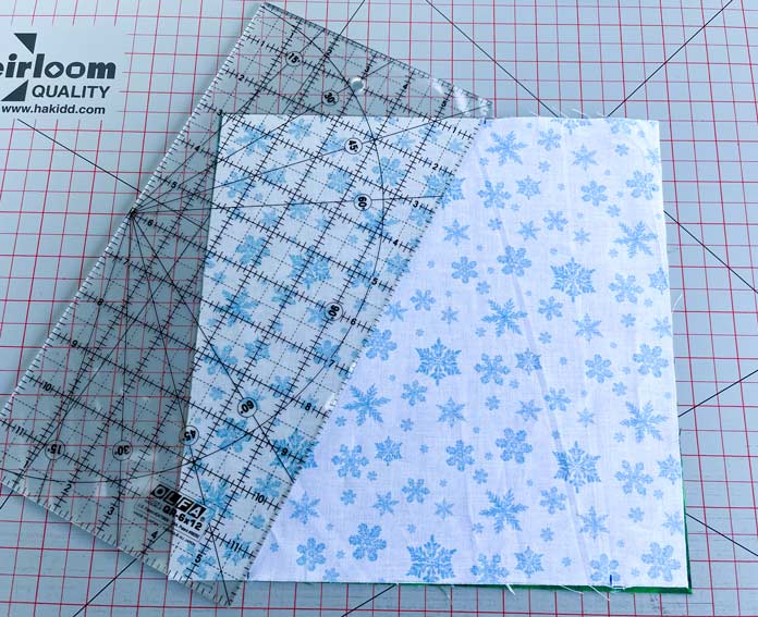 On a gray cutting mat with red grid lines, a quilting ruler is placed diagonally on a square piece of white and blue fabric going from one mark to another; OLFA 6″ x 12″ Frosted Acrylic Ruler, Heirloom 24″ x 36″ Double Sided Cutting Mat