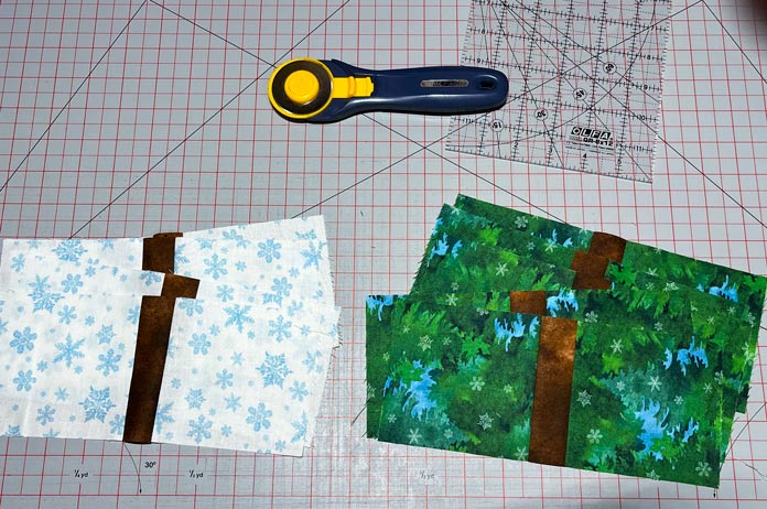 2 stacks of rectangular shape blocks, one with a brown strip and snowflake fabric on either side and one with a brown strip and green fabric on either side, lay on a gray cutting mat with red gridlines. There’s a rotary cutter and quilting ruler above the fabric blocks