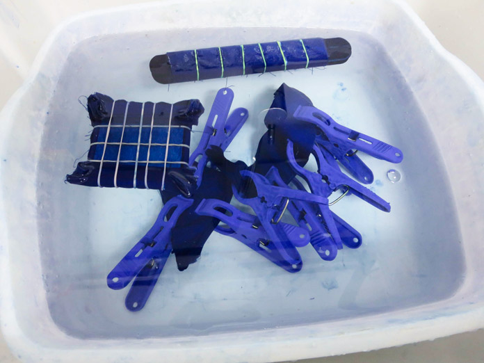 Dyed fabric wrapped using rubber bands, craft sticks and clothes pins in the ColorStay Dye Fixative solution in a white basin; Rit Indigo Shibori Tie Dye Kit, Rit Indigo All-purpose Dye, Rit ColorStay Dye Fixative