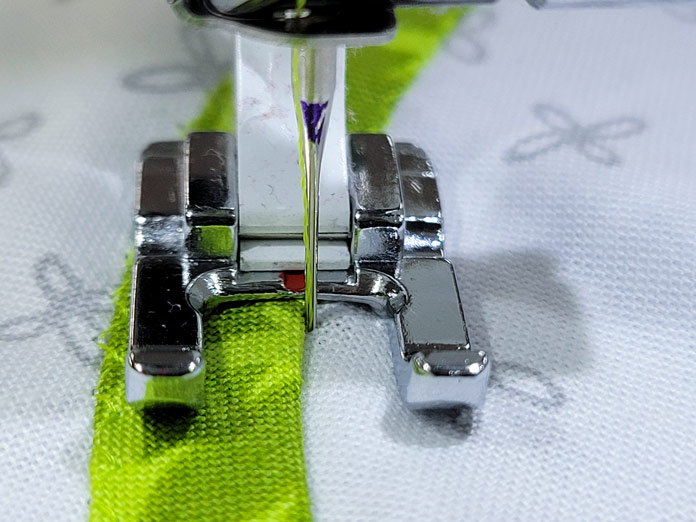 A metal presser foot with a strip of green fabric on a white background; Husqvarna Viking Tribute 150C