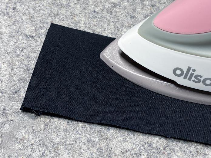 A pink mini OLISO M2Pro iron is shown pressing the folded edge of a black rectangle of fabric on top of a gray wool pressing mat.