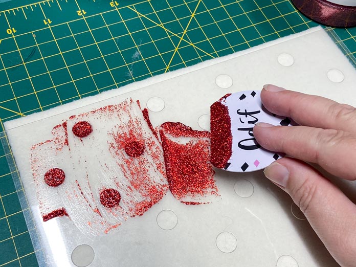 A hand is shown holding a white plastic applicator which has red glitter gel on its straight edge. The gel has been spread on a clear plastic polka dot stencil stuck to a rectangle of cream-colored felted wool. In the background is a green cutting mat.