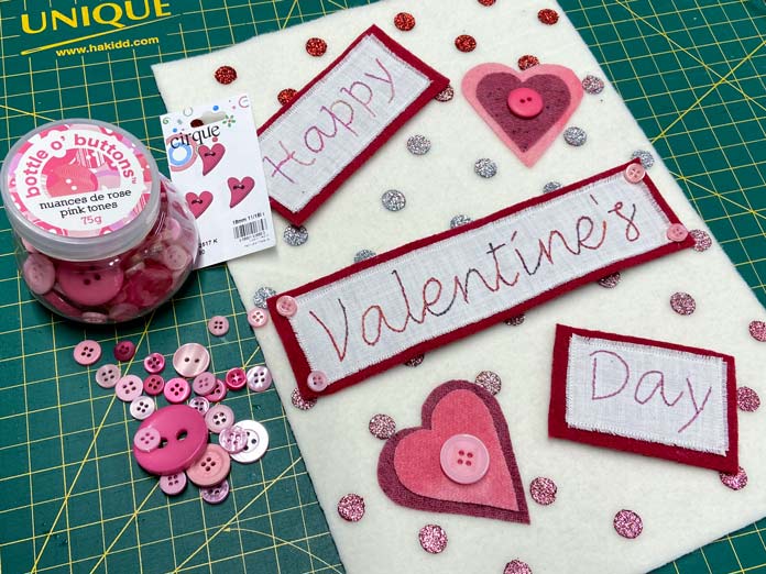 A bottle of pink buttons along with some loose pink buttons sit on a green cutting mat beside a cream, red and pink wall banner that says Happy Valentine’s Day. CRAFTING ESSENTIALS Bottle of Buttons - Pink Tones - 75g (2.6oz), HEATNBOND EZ Print Feather Lite 10 pcs - 22 x 28cm (81⁄2″ x 11″), SULKY Cotton Petites 6 Spool Thread Set - Rosewood Manor Assortment, UNIQUE Hook & Loop Sew-On Tape - 19mm x 10m (3⁄4″ x 11yd) - Black, OLFA RTY-2/DX - Deluxe Ergonomic Handle Rotary Cutter 45mm, OMNIGRID Ruler - 6″ x 12″, ELAN Novelty 2-Hole Button - Rose - 18mm (3⁄4″) - Heart - 3 count, UNIQUE SEWING Chenille Needles - sizes 18/24 - 6pcs, ODIF OdiShine Glitter Gel, DMC Embroidery Tracing/Transfer Paper, UNIQUE QUILTING Clever Clips Small - 12 pcs, free tutorial.