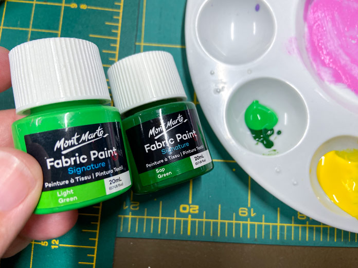 A hand holding two different green and black jars of paint is shown beside a round, white, plastic artist’s palette. There are different colors of paint on the palette; Mont Marte Round Plastic Palette 6¾”, Mont Marte Signature Fabric Paints