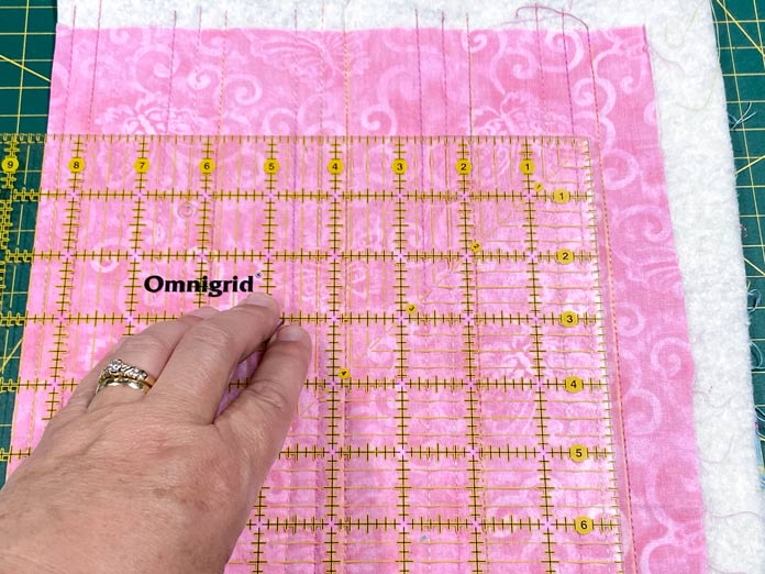A hand is shown holding an Omnigrid ruler on top of a small pink quilt. The batting in the quilt is visible past the edges of the pink fabric and everything is sitting on a green cutting mat.