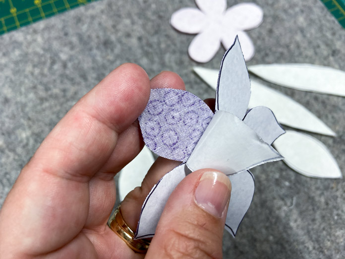 A hand is shown peeling away the paper backing from a purple fabric flower. A gray wool pressing mat is shown in the background; UNIQUE Quilting Wool Pressing Mat - 14″ x 14″, HeatnBond EZ Print Feather Lite 10 pcs - 22 x 28cm (8½″ x 11″)