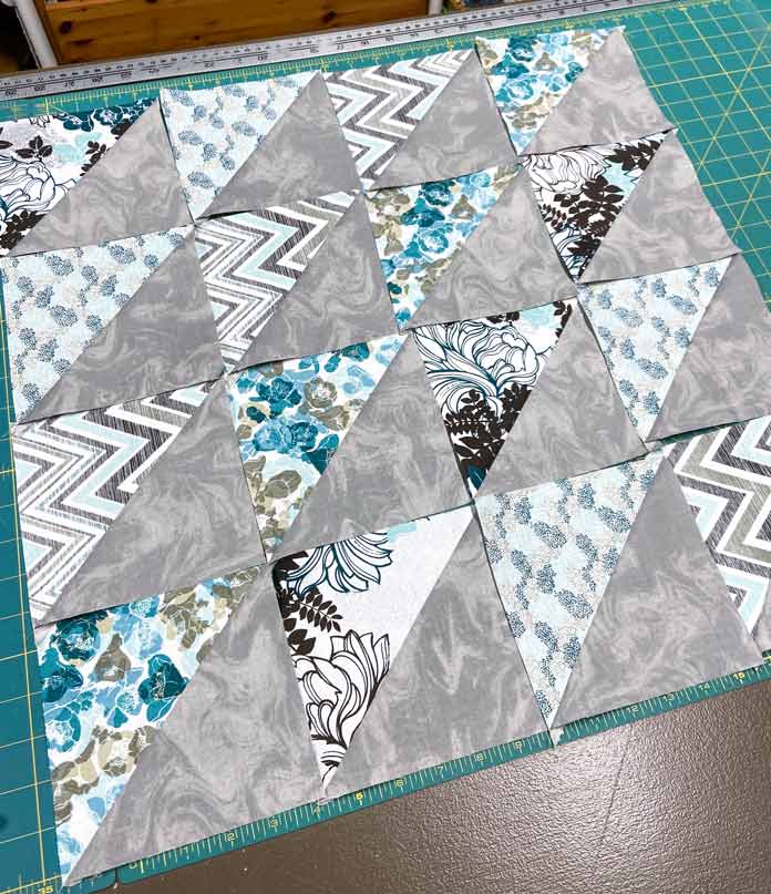 Sixteen grey, teal, turquoise and white half square triangle units are arranged into a square on top of a green cutting mat. Omnigrid Triangle Ruler for Half Square Triangles, Fabric Creations Trendy Neutral Fabric collection, Fabric Creations Fabric Palette collection, Clover Hot Hemmer, UNIQUE quilting Clever Clips, UNIQUE Clear Grip, Oliso Pro TG1600 Smart Iron, Fairfield Crafter’s Choice Pillow Forms, StitchnSew EZ-Print Quilt Block Sheets