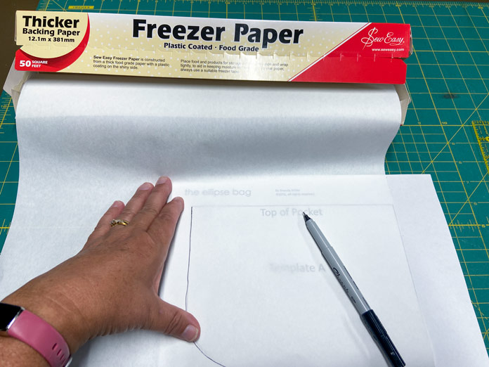 A hand is shown tracing a template with a black marker onto the paper side of a roll of freezer paper. The box of freezer paper is in the background, and everything sits on a green cutting mat.