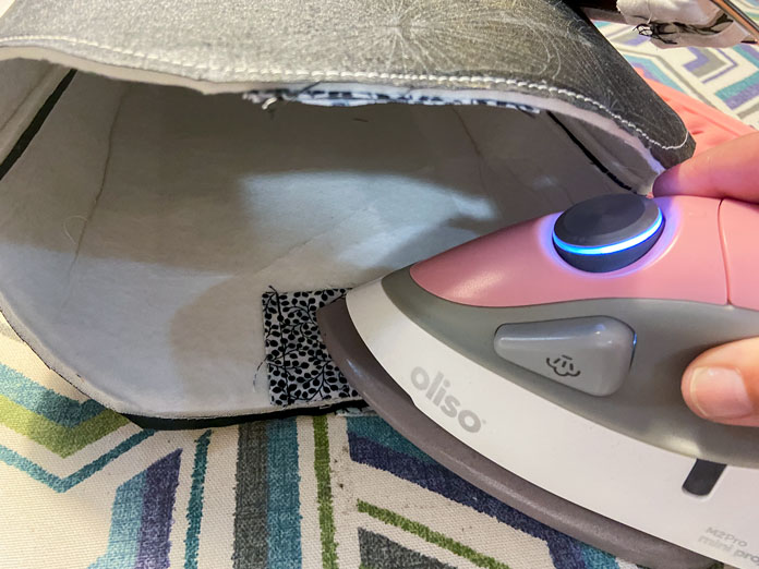 A small pink iron is shown ironing a black and white pouch to the inside of a tote bag. A multicolored ironing board is in the background.