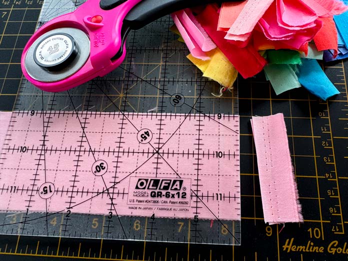 An OLFA 6” x 12” quilting ruler and a pink OLFA rotary cutter are being used to cut the selvage edge off a pink strip of fabric with a pile of brightly colored selvages from other strips off to the side. Sewing Kit, Hemline Gold Quilters 4-in-1 Multi-Mat (30cm x 24cm), Hemline GoldQuilters Clips (Pack of 30), Hemlione Gold Retractable Tape Measure - 150cm/60, oliso M3Pro Project Iron – Coral, OLFA 45mm RTY-2/DX/MAG Ergonomic Rotary Cutter – Magenta, OLFA RM-MG - 24″ x 36″ Double Sided Rotary Mat, UNIQUE Sewing Tools Holder, OLFA QR-6x12 - 6″ x 12″ Frosted Acrylic Ruler, OLFA QR-6x24 - 6″ x 24″ Frosted Acrylic Ruler, Fairfield Quilter's 80/20 Quilt Batting - 229 x 274cm (90″ x 108″)