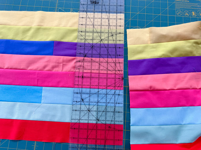 A 54” section is removed from a long set of 8 pieced multicolor fabric strips using a frosted OLFA ruler on a green cutting mat. Hemline Gold Multi-Use Craft Bag (28.5cm x 36.5cm x 17.8cm), Hemline Gold Sewing Kit, Hemline Gold Quilters 4-in-1 Multi-Mat (30cm x 24cm), Hemline Gold Quilters Clips (Pack of 30), Hemlione Gold Retractable Tape Measure - 150cm/60, Oliso M3Pro Project Iron – Coral, OLFA 45mm RTY-2/DX/MAG Ergonomic Rotary Cutter – Magenta, OLFA RM-MG - 24″ x 36″ Double Sided Rotary Mat, UNIQUE Sewing Tools Holder, OLFA QR-6x12 - 6″ x 12″ Frosted Acrylic Ruler, OLFA QR-6x24 - 6″ x 24″ Frosted Acrylic Ruler, Fairfield Quilter's 80/20 Quilt Batting - 229 x 274cm (90″ x 108″)