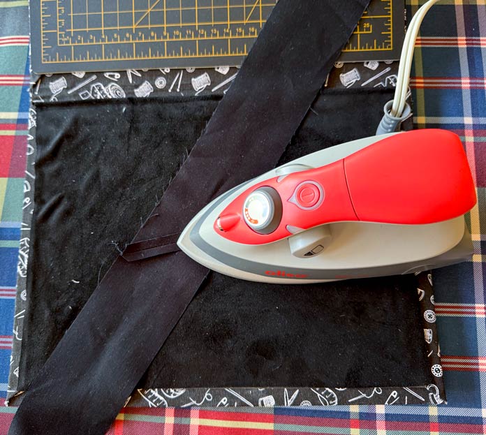 A seam sewn in two strips of black fabric is being pressed open using a coral-colored oliso M3Pro project iron on top of a black pressing mat with a plaid fabric in the background. Hemline Gold Multi-Use Craft Bag (28.5cm x 36.5cm x 17.8cm), Hemline Gold Sewing Kit, Hemline Gold Quilters 4-in-1 Multi-Mat (30cm x 24cm), Hemline Gold Quilters Clips (Pack of 30), Hemline Gold Retractable Tape Measure - 150cm/60, oliso M3Pro Project Iron – Coral, OLFA 45mm RTY-2/DX/MAG Ergonomic Rotary Cutter – Magenta, OLFA RM-MG - 24″ x 36″ Double Sided Rotary Mat, UNIQUE Sewing Tools Holder, OLFA QR-6x12 - 6″ x 12″ Frosted Acrylic Ruler, OLFA QR-6x24 - 6″ x 24″ Frosted Acrylic Ruler, Fairfield Quilter's 80/20 Quilt Batting - 229 x 274cm (90″ x 108″) 