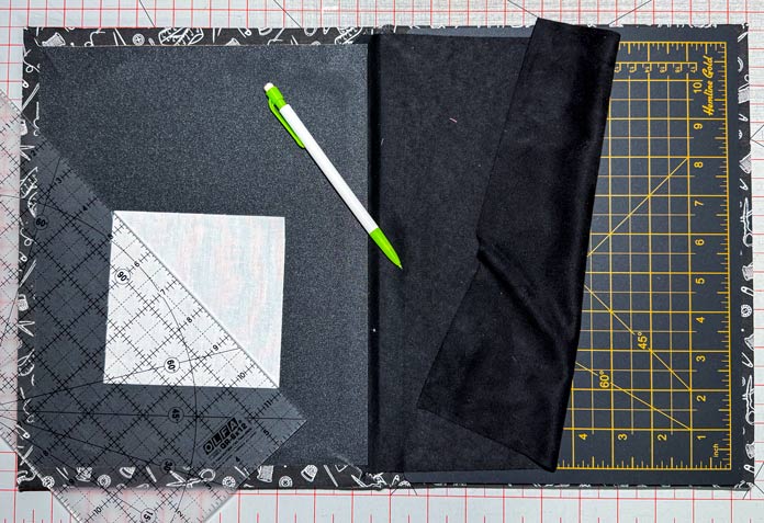 A white square of fabric with a quilting ruler placed diagonally on top is placed on a sheet of fine gray sandpaper that is part of a Hemline Gold Quilters 4-in-1 Multi-Mat. A green and white marking pen is laid to the side while a gray cutting mat is revealed beside the sandpaper by folding back a black, nonslip fabric. A light gray cutting mat is in the background. Hemline Gold Multi-Use Craft Bag (28.5cm x 36.5cm x 17.8cm), Hemline Gold Sewing Kit, Hemline Gold Quilters 4-in-1 Multi-Mat (30cm x 24cm), Hemline Gold Quilters Clips (Pack of 30), Hemline Gold Retractable Tape Measure - 150cm/60, oliso M3Pro Project Iron – Coral, OLFA 45mm RTY-2/DX/MAG Ergonomic Rotary Cutter – Magenta, OLFA RM-MG - 24″ x 36″ Double Sided Rotary Mat, UNIQUE Sewing Tools Holder, OLFA QR-6x12 - 6″ x 12″ Frosted Acrylic Ruler, OLFA QR-6x24 - 6″ x 24″ Frosted Acrylic Ruler, Fairfield Quilter's 80/20 Quilt Batting - 229 x 274cm (90″ x 108″) 