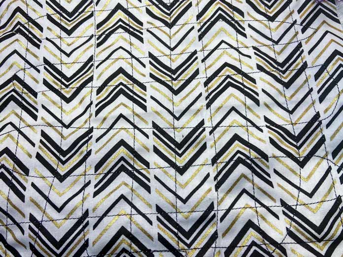 A closeup of a fabric with a black and gold chevron design that has horizontal and vertical lines of black stitching. Oliso M2Pro Mini Project Iron, FAIRFIELD Low-Loft Quilt Batting - Crib - 114 x 152cm (45″ x 60″), UNIQUE QUILTING Clever Clips, OLFA CHN-1 - Chenille / Textile Cutter, UNIQUE CREATIV Trim Pack - Black & White, FABRIC CREATIONS Burlap Fabric - Oyster - 45 x 61cm (18″ x 24″), FABRIC CREATIONS Faux Fur Fabric - Zebra - 45 x 76cm (18″ x 30″), UNIQUE SEWING Double Adjust Parachute Buckle - Plastic - 25mm (1″) White, CRAFTING ESSENTIALS Bottle of Buttons - Pink Tones - 75g (2.6oz), UNIQUE SEWING Invisible Magnetic Snaps - Sew-On - 12mm (1⁄2″) - 2 sets, UNIQUE QUILTING Stitch-N-Steam - 157 x 45cm (62″ x 18″), FABRIC CREATIONS Fabric Bundle (5pcs) - Metallic Gold - 45 x 53cm (18in x 21in), Oliso Pro TG1600 Pro Plus Smart Iron, UNIQUE QUILTING Clever Clips Large - 12pcs, sewing tutorial