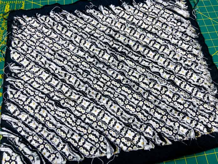 A rectangle of black and white chenille fabric sits on top of a green cutting mat. Oliso M2Pro Mini Project Iron, FAIRFIELD Low-Loft Quilt Batting - Crib - 114 x 152cm (45″ x 60″), UNIQUE QUILTING Clever Clips, OLFA CHN-1 - Chenille / Textile Cutter, UNIQUE CREATIV Trim Pack - Black & White, FABRIC CREATIONS Burlap Fabric - Oyster - 45 x 61cm (18″ x 24″), FABRIC CREATIONS Faux Fur Fabric - Zebra - 45 x 76cm (18″ x 30″), UNIQUE SEWING Double Adjust Parachute Buckle - Plastic - 25mm (1″) White, CRAFTING ESSENTIALS Bottle of Buttons - Pink Tones - 75g (2.6oz), UNIQUE SEWING Invisible Magnetic Snaps - Sew-On - 12mm (1⁄2″) - 2 sets, UNIQUE QUILTING Stitch-N-Steam - 157 x 45cm (62″ x 18″), FABRIC CREATIONS Fabric Bundle (5pcs) - Metallic Gold - 45 x 53cm (18in x 21in), Oliso Pro TG1600 Pro Plus Smart Iron, UNIQUE QUILTING Clever Clips Large - 12pcs, UNIQUE SEWING 2-in-1 Dual-Tip Wash-out/Air Erasable Marking Pen - Fine,OMNIGRID Ruler - 121⁄2″ x 121⁄2″ (31.75 x 31.75cm), sewing tutorial