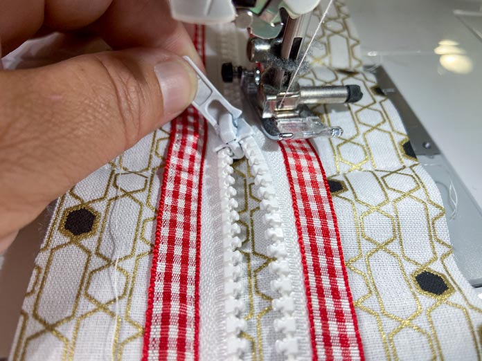 A sewing machine stitches along the edge of a red gingham ribbon and a white zipper while a hand holds the zipper pull away from the foot of the sewing machine. UNIQUE SEWING Self-Gripping Fastener Strip - 25cm (10″) - White, UNIQUE Webbing 25mm x 10m (1″ x 11yd) - White, ELAN Trench Buckle - 45mm (13⁄4″) - Brown, UNIQUE CREATIV Trim Pack - Black & White, UNIQUE SEWING Invisible Magnetic Snaps - Sew-On - 12mm (1⁄2″) - 2 sets,  SOFTKUT Pinking Shears (Scissors) - 81⁄2″ (21.6cm), COSTUMAKERS General Purpose Closed End Zipper 20cm (8″) - White - 1700, UNIQUE CREATIV Trim Pack - Red & Green, OLFA RTY-2/DX - Deluxe Ergonomic Handle Rotary Cutter 45mm, UNIQUE QUILTING Clever Clips Small - 12 pcs, UNIQUE SEWING Double Adjust Parachute Buckle - Plastic - 25mm (1″) - White, CRAFTING ESSENTIALS Bottle of Buttons - Pink Tones - 75g (2.6oz), UNIQUE Braided Elastic 3mm x 3.7m - White, free tutorial