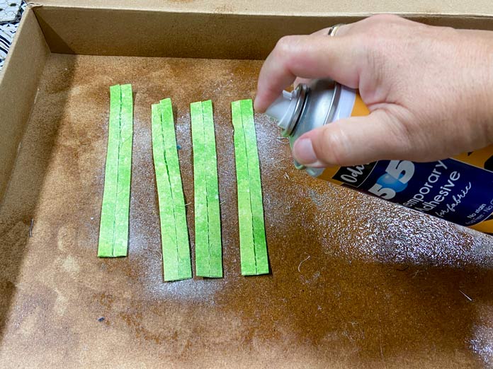 A hand holds a yellow and blue can of adhesive spray over 4 strips of green fabric. A brown cardboard box with a shiny surface can be seen in the background. UNIQUE SEWING Invisible Magnetic Snaps - Sew-On - 12mm (1⁄2″) - 2 sets, COSTUMAKERS General Purpose Closed End Zipper 20cm (8″) - White - 1700, UNIQUE SEWING Double Adjust Parachute Buckle - Plastic - 25mm (1″) – White, UNIQUE QUILTING Clever Clips Small - 12 pcs, CLOVER 7812 Roll & Press, UNIQUE QUILTING Wool Pressing Mat - 18″ x 24″ - Grey, OLISO M2Pro Mini Project Iron with Solemate, CLOVER 240 - Fork Pins - .56mm, CLOVER 464/18 - Bias Tape Maker - 18mm (3⁄4″), Odif 505 Temporary Quilt Basting Adhesive Fabric Spray - 156g, FAIRFIELD Low-Loft® Quilt Batting - Crib - 114 x 152cm (45″ x 60″), free tutorial