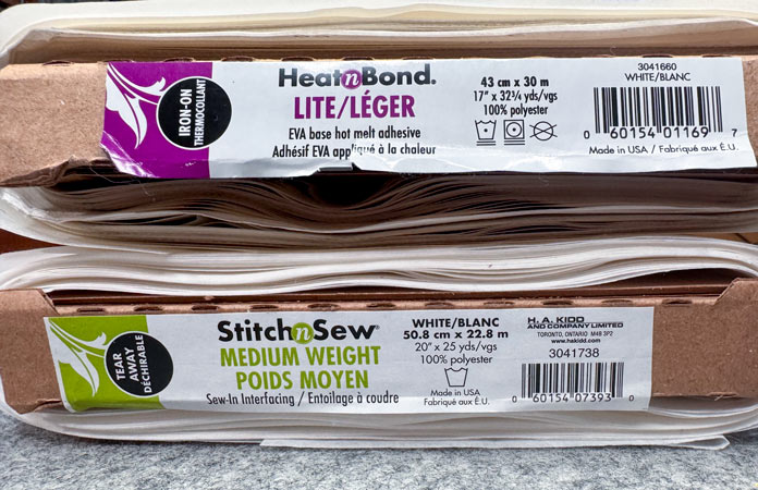 A bolt of HeatnBond Lite adhesive and a bolt of Stitch n Sew medium weight tear away on top of a gray pressing mat; Gütermann Dekor Metallic Thread 200m – Gold, Gütermann Dekor Metallic Thread 200m - Col. 41, Gütermann Dekor Metallic Thread 200m – Red, Gütermann Dekor Metallic Thread 200m - Christmas Green, Gütermann Dekor Metallic Thread 200m - Col. 1005, Gütermann Denim Thread 100m – Black, UNIQUE Baby Rick Rack 3mm x 3.7m – Scarlet, UNIQUE Medium Rick Rack 6mm x 2.3m - Emerald Green, UNIQUE Jumbo Rick Rack 10mm x 2.3m - Metallic Gold, SCHMETZ #1743 Metallic Needles Carded - 80/12 - 5 count, SCHMETZ #1752 Metallic Needles Carded - 90/14 - 5 count, LDH 8½“ Craft Scissors – Purple, HeatnBond Lite Iron-On Adhesive - 43cm x 30m (17″ x 32 ¾yds) bolt, HeatnBond Stitch n Sew Tear-Away Stabilizer - 50.8cm x 22.8m (20″ x 25yds), oliso M3Pro Project Iron – Coral, OLFA 45mm RTY-2/DX/MAG Ergonomic Rotary Cutter – Magenta, UNIQUE Quilting Wool Pressing Mat - 18″ x 24″ - Grey