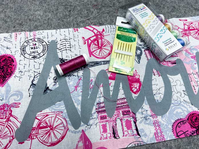 A white package of thread, a dark pink spool of thread and a yellow package of chenille needles sit on top of the pink, black and white fabric that has the appliqued word on it. A mottled gray wool pressing mat is in the background.