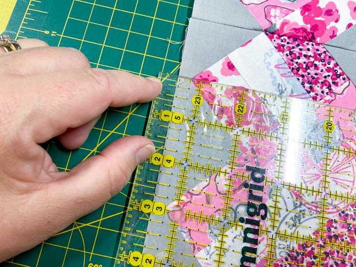 A hand uses a rotary cutting ruler with a yellow grid to measure the length of a pink, white and gray quilt. A green cutting mat is in the background. 