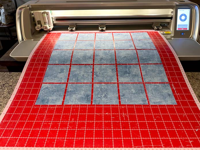 A picture of blue fabric cut into squares on a red mat coming out of the SINGER MOMENTO 24 digital cutter machine; quilting, digital quilting, fabric cutting, SINGER MOMENTO 24, mySewnet Crafting Software, quilt design, quilting tutorial, precision cutting, beginner quilting, quilt planning