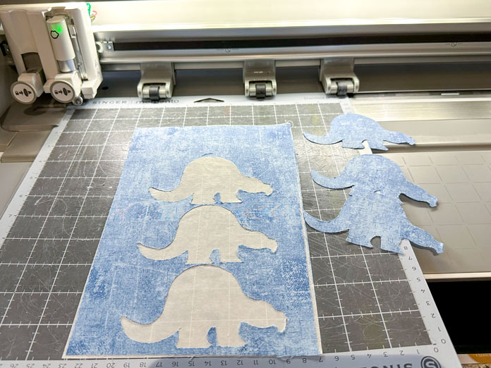 A picture of the SINGER MOMENTUM 24 with the grey standard mat and the three right-facing dinosaur body applique pieces cut out; quilting, embroidery, applique, mySewnet Silver, digital quilting, SINGER MOMENTO 24, PFAFF creative 350, fabric cutting, quilt design, beginner quilting