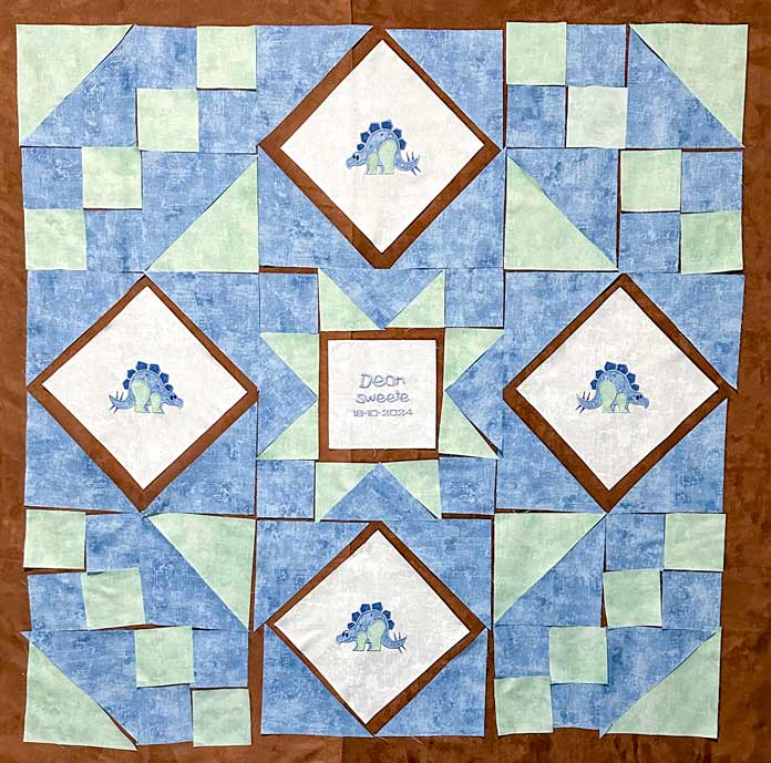 A brown design wall with blue and green fabric pieces arranged in the Road to Oklahoma pattern in the 4 corners, the Ohio Star block  in the center with a white embroidered block in the center of it (with a name and birthdate embroidered on it), and lastly 4 Almond  (square in a square) blocks at the NS, EW positions with the embroidered blue dinosaur on white fabric as the center; mySewnet Quilting Software, PFAFF creative expect 350, mini quilt tutorial, beginner quilting, machine embroidery techniques, applique embroidery, sewing and quilting, quilt assembly, machine embroidery tips, DIY quilt projects, PFAFF, sewing machine reviews, quilting tutorials, free quilting patterns