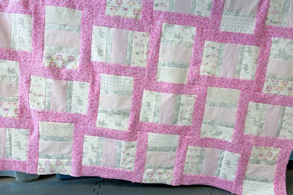 A pink, gray and white quilt hangs off one rail of a longarm quilting machine. A grey mat is on the floor beneath.