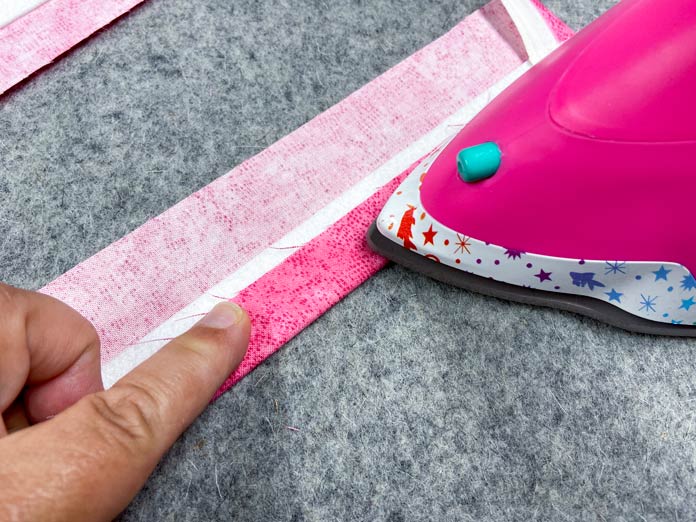 A hand is holding down the folded edge of a strip of pink fabric, so that a pink iron can press the fabric to the white interfacing strip underneath. Everything sits on a grey pressing mat.