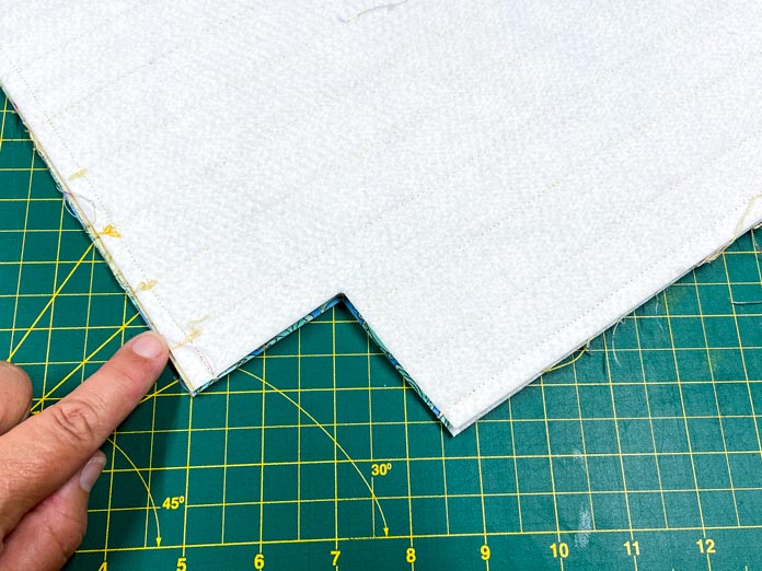 A finger points to the stitching that is holding together two panels for a tote bag. The stitching is visible along the sides and bottom. There is a square in the corner that has been cut out. A green cutting mat is in the background. 