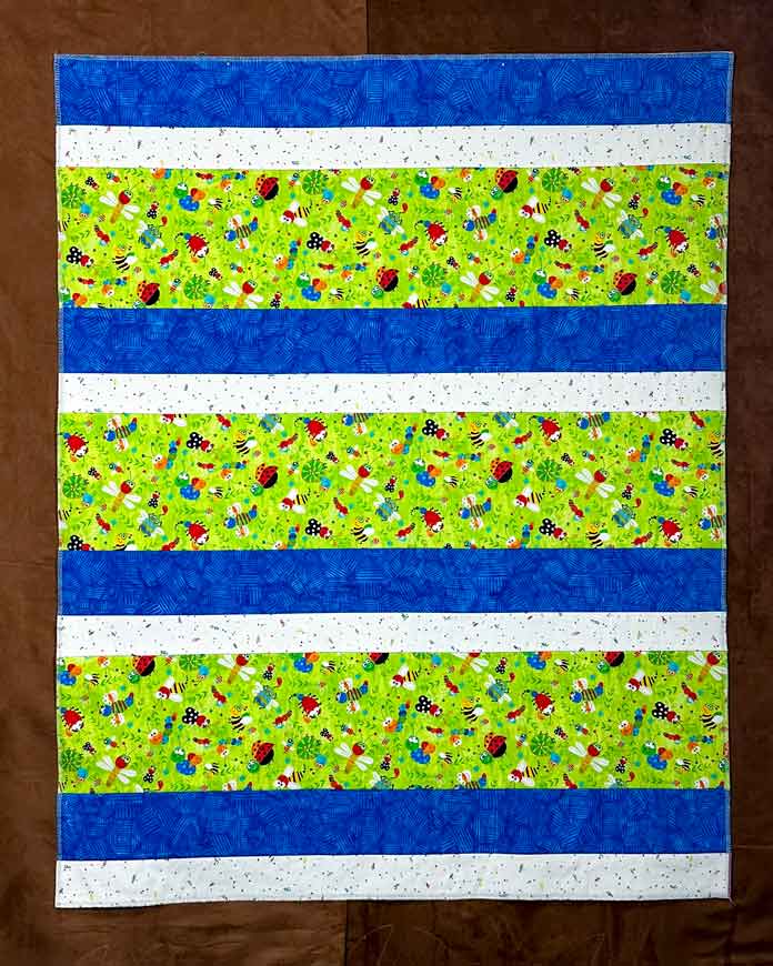 White print, blue batik, green bug print, in (2.5”,4”,8.5”) finished strips repeated 3X, ending on an extra white print and blue batik strip. Serged top and bottom, selvedges removed left and right, and these sides serged, ready now for binding.