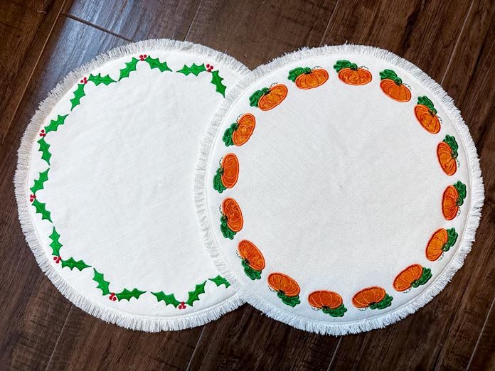 2 cream circle placemats with frayed edge and embroidered pumpkins and leaves on one and embroidered in a circle holly leaves and holly red berries.