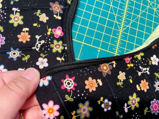 A hand holds a purse panel with a zipper sewn into the top edge; a second panel lies on a green cutting board in the background.