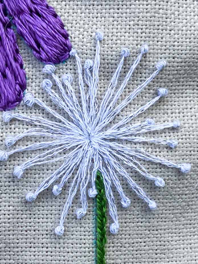 A close-up photo shows how pistil stitches are embroidered using sparkly white DMC Mouline Etoile floss. The stitches all start in the middle of a flower and radiate outward to create a dandelion shape. There is a green stem embroidered beneath it and in the top left corner you can see a portion of the purple flower. All are shown on beige Charles Craft Monaco Needlework Fabric.