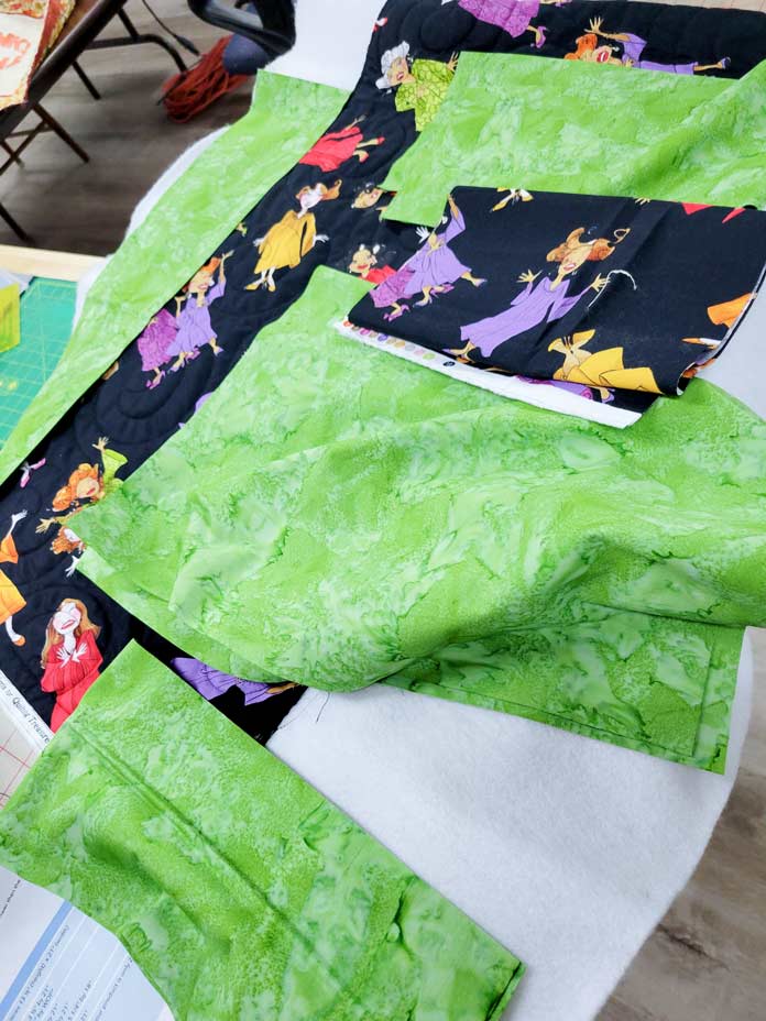 Several green fabric pieces and black fabric with ladies dressed in colorful outfits; Husqvarna Viking Designer Ruby 90 sewing and embroidery machine, Inspira Fusible Fleece, Omnigrip 20½" square ruler, Singer 20 Steam Garment Press