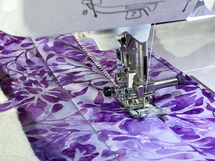 A sewing machine is shown sewing a zigzag stitch along the edge of a purple floral batik fabric. Brother NQ900 sewing machine, UNIQUE quilting Clever Clips, UNIQUE Medium Rick Rack