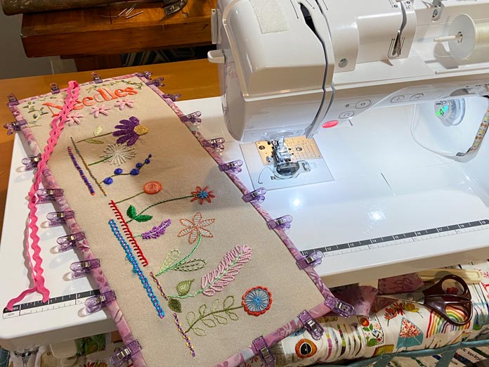 A beige fabric needle roll with the multicolored embroidery is shown on the extension table of the NQ900 sewing machine, with purple plastic clips in place all around holding the pink batik binding secure.