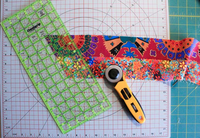 Two strips of fabric sewn together on a cutting mat and next to a quilting ruler and rotary cutter; Komfort Kut 360° Rotating Cutting Mat - 18" x 18", Omnigrip Ruler - 6″ x 12″, OLFA Quick Change Rotary Cutter
