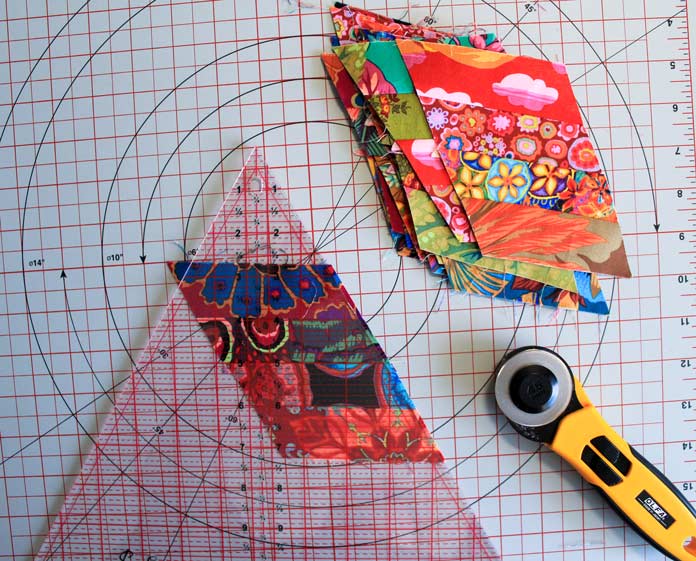 Trimming off the excess fabric on the edge of a pieced unit using a triangle ruler, rotary cutter, and cutting mat; Komfort Kut 360° Rotating Cutting Mat - 18" x 18", Sew Easy Triangle Ruler 60°, OLFA Quick Change Rotary Cutter