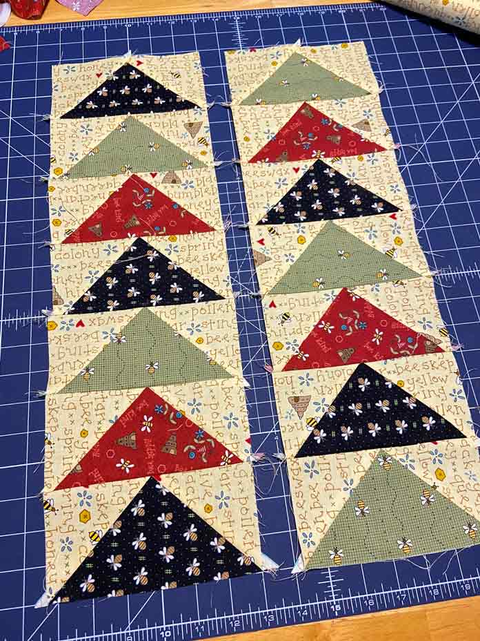 Two rows of seven flying geese blocks in red, black green and yellow lay on a dark blue cutting mat; OLFA Double Sided Cutting Mat (navy blue)