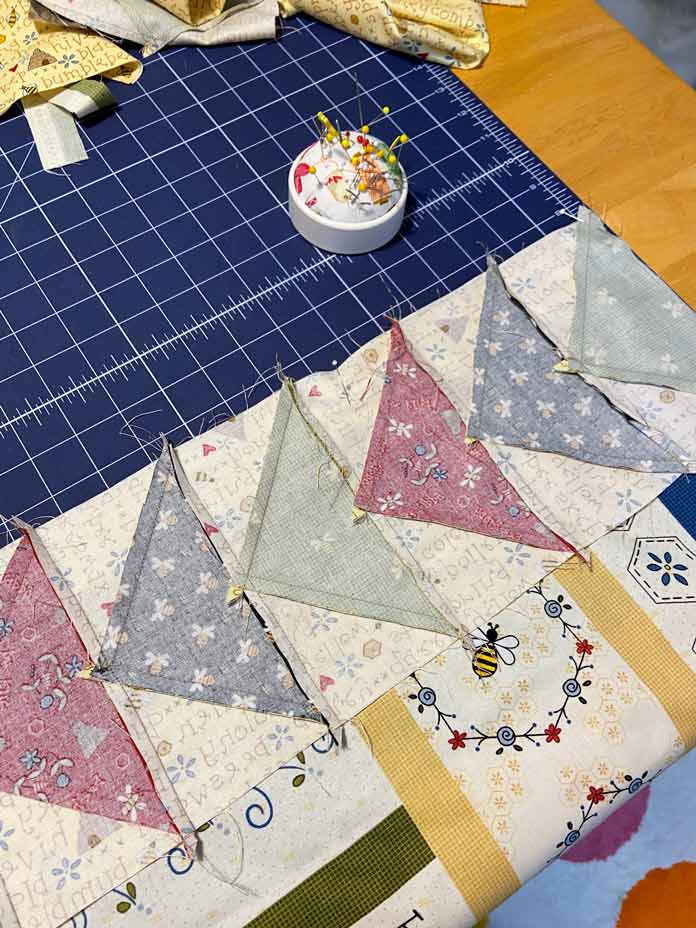 The flying geese border made of black, green and red flying geese is shown pinned right side together with the bee-themed panel. A blue cutting mat and a small round pin cushion are in the background; OLFA Double Sided Cutting Mat (navy blue)