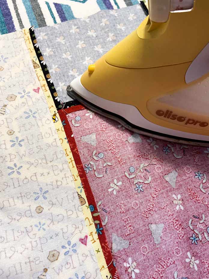 A yellow Oliso Pro iron is shown pressing the seam between a strip of multicolored fabric and a larger piece of yellow fabric on top of a white, teal, blue and gray geometric ironing board.