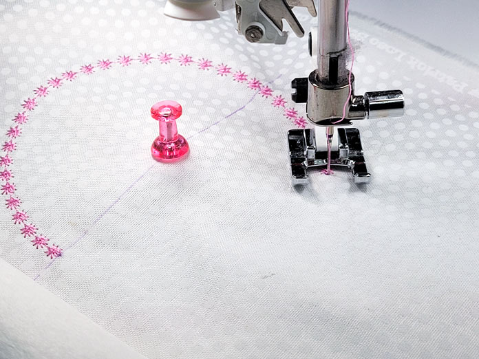 A pink push pin, a partial circle stitched in pink thread, and a sewing machine presser foot; Husqvarna Viking Designer EPIC 2
