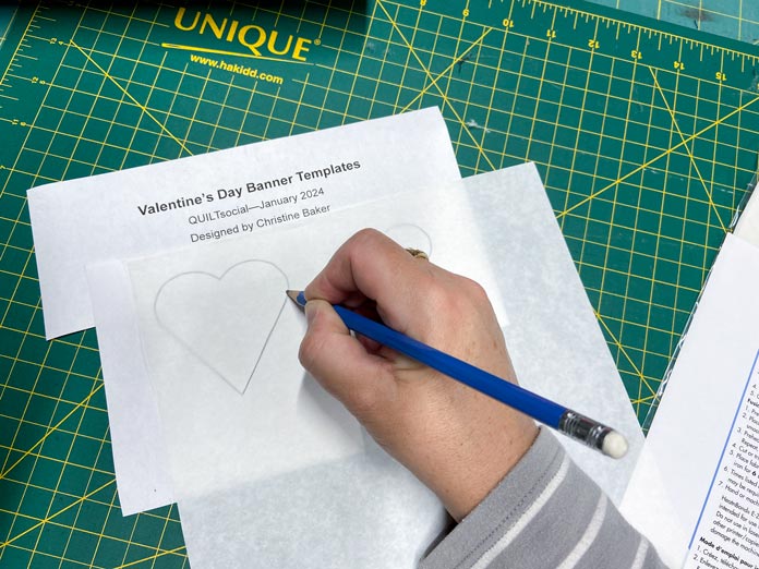 A hand traces a heart shaped template on to the paper side of one piece of HeatnBond EZ Print Feather Lite iron-on adhesive using a blue pencil. In the background is a green cutting mat. 