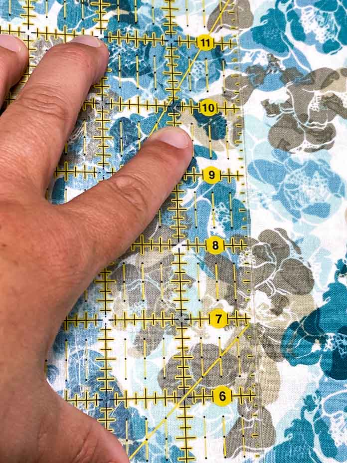 A hand holds a yellow gridded quilting ruler with a floral fabric behind it. UNIQUE Clear Grip, UNIQUE quilting Ruler/Template Grips, Omnigrid Ruler, Omnigrid Triangle Ruler, Fabric Creations Fabric Palette Pre-Cut Fabrics, Fairfield Crafter’s Choice Pillow Form