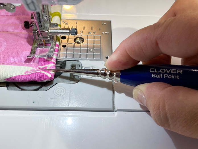 A hand is shown holding a blue handled awl which is being used to hold down the corner of the binding on a quilt while the sewing machine is topstitching; Clover 876 – Ball Point Awl 