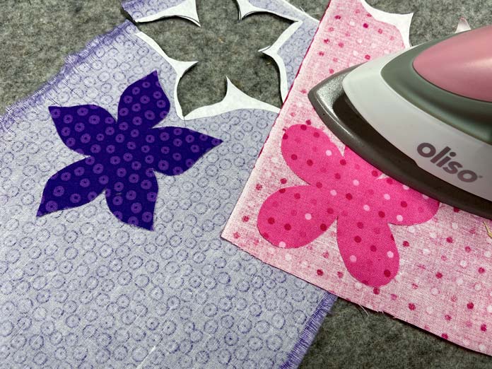 A pink Oliso Mini Project iron is shown ironing a pink fabric flower shape onto the back of a pink fabric. A purple flower shape and a gray wool pressing mat are shown in the background; UNIQUE Quilting Wool Pressing Mat - 14″ x 14″, HeatnBond EZ Print Feather Lite 10 pcs - 22 x 28cm (8½″ x 11″)