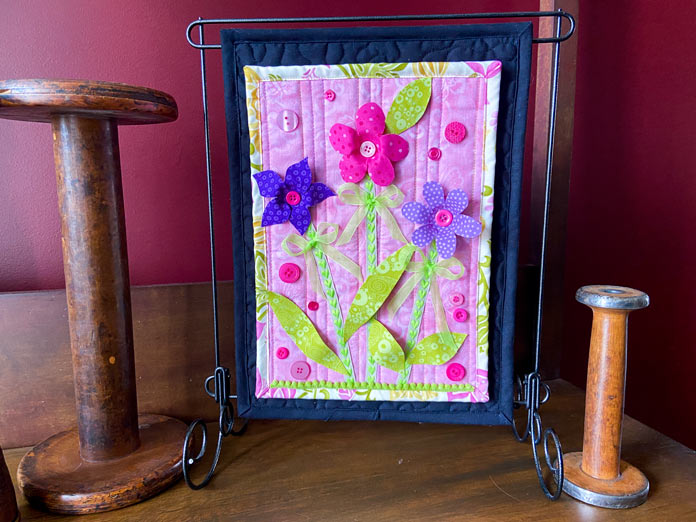 A small pink quilt with three-dimensional pink and purple flowers and green leaves is shown hanging on a metal stand on top of a wooden table. A couple of antique wooden spools are sitting beside it.