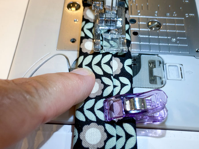 A hand is shown folding under the ends of a fabric strap. The white extension table of a sewing machine is shown in the background.