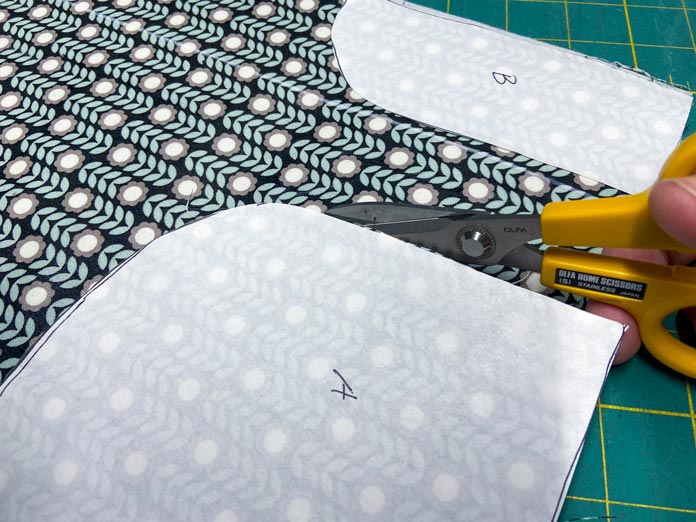 A hand is shown cutting along the edge of a template with yellow scissors. In the background is a green cutting mat.